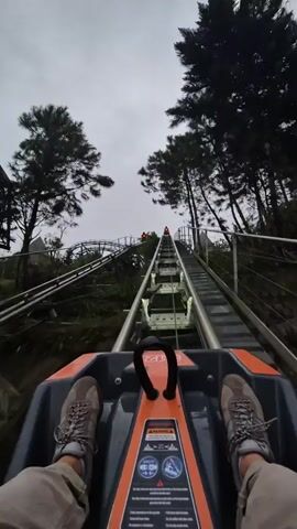 The first-person point-of-view shows a person riding an alpine coaster uphill on a track. The person wears brown hiking boots and tan pants. The coaster seat is gray and orange, with a black hand grip. The metal track leads upward between green trees under a gray sky. Another coaster is ahead on the track. A resort building is visible in the background on the right.