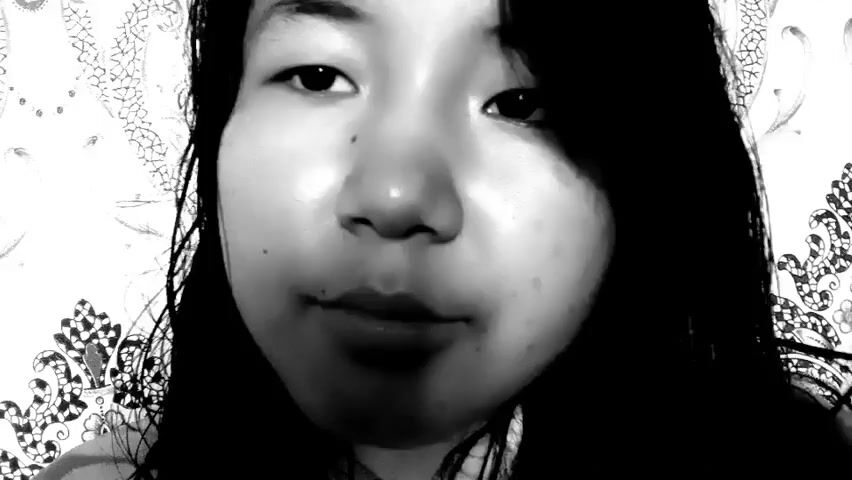 This is a black and white video recording of a young Asian woman. She is positioned close to the camera, occupying most of the frame. The background consists of a patterned wallpaper, featuring ornate, repeating designs. The lighting is somewhat dramatic, casting shadows and highlighting the woman's face, giving it a glossy appearance.

The woman's hair is dark and falls around her face and shoulders. She is looking directly at the camera and her expression shifts throughout the recording. At times she appears to be talking, with subtle movements of her lips. She also interacts with her hair, touching it and adjusting it. Her emotions seem to change from thoughtful or perhaps slightly pensive, to a more engaged and smiling expression. There are moments where she seems to be making a playful or charming gesture.

The recording appears to be a selfie-style video, possibly taken indoors in a residential setting. There are no obvious objects other than the wallpaper and the woman herself. The time of day and weather are not discernible. No text is visible in the media file. The overall impression is of a personal, candid moment captured on video.