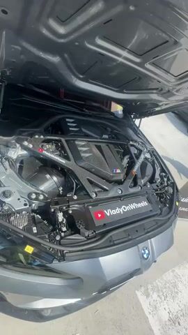 A gray BMW M4 is parked outside with its hood open, revealing the engine. A person's arm is visible on the left side of the screen, partially obscuring the car. The car is on a paved surface, possibly a parking lot or a race track, with a white tent in the background. The car has custom rims and visible brake calipers. The video pans across the engine bay and then focuses on the front wheel of the car. A YouTube logo with "VladyOnWheels" is visible on the engine cover.
