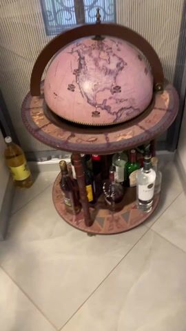The video shows a pink globe bar stand. The globe has brown trim and a brown semi-circular handle around the top. The surface of the globe depicts land masses. The person in the video opens the globe at the 0:03 mark to reveal the inside. The inside has illustrations of birds, ships, and other designs in black, white, and brown colors. Below the globe is a circular shelf holding various bottles of liquor. The surface of this shelf is decorated with brown and purple patterns. The globe stand is sitting on a light-colored tiled floor near a striped curtain.