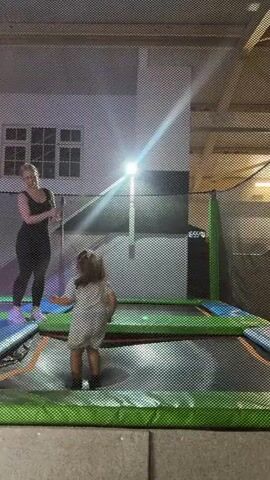 Two young people are on a trampoline. One person in a white outfit is mid-flip, upside down with their legs in the air. The other person, dressed in black, stands on the side of the trampoline and appears to be holding a hula hoop. There is another child in the background on a different trampoline. The scene is indoors, possibly a trampoline park, with netting surrounding the trampoline area. The lighting is bright, with a spotlight shining down.