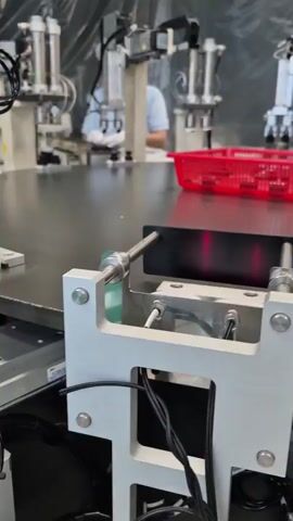 The video shows an indoor industrial setting, likely a factory floor. In the foreground, a close-up view focuses on metallic robotic machinery with visible wiring and electrical components. The machinery appears to be part of an automated assembly or processing line. In the background, a person wearing a white cap and light-colored clothing is visible, interacting with other machinery. A red plastic basket containing small components is also in the background, suggesting a workflow or inventory management system. The overall environment is clean and organized, typical of a manufacturing facility. The camera pans across the machinery, highlighting its functional aspects. The audio contains some muffled speech in a language other than English and the ambient sounds of machinery operating.