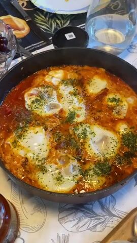 A close-up, high-angle shot captures a skillet filled with shakshuka, a dish of eggs poached in a tomato and pepper sauce. The eggs are cooked to varying degrees, with some having fully set whites and others still showing a liquid yolk. The dish is garnished with chopped parsley. The skillet sits on a table covered with a patterned tablecloth. In the background, a glass of water, a bowl of cherries, and some other dining items are visible. The lighting suggests an indoor setting, likely a dining room or kitchen.