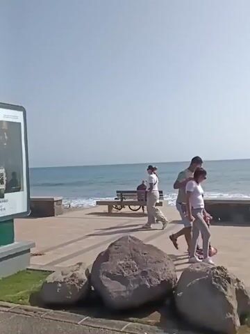 The video shows a seaside promenade on a bright, sunny day. The sky is clear blue, and the ocean is visible with small waves. On the promenade, people are walking leisurely, some are in groups. A tall, cylindrical lighthouse stands in the background, serving as a prominent landmark. Closer to the camera, there's a billboard advertising a watch. A man is sitting on the ground near the billboard, seemingly organizing his belongings. There are also benches and decorative boulders along the promenade. The ground is paved, and there's some landscaping with grass and plants. A restaurant with outdoor seating is visible.