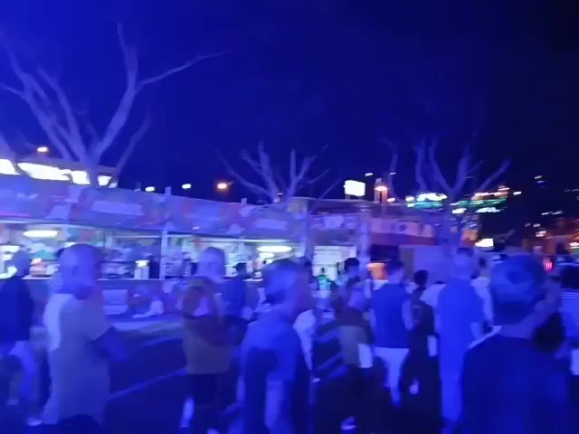 The video shows a crowd of people at an outdoor event at night. The event appears to be a concert or festival, as there is a large screen displaying a live video feed of a performer. There are also food and drink vendors set up in the background. The scene is lit with blue and green lighting. Most of the people are facing the stage and seem to be enjoying the show.