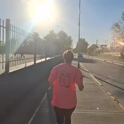 A person is seen from behind, running or jogging on a paved sidewalk in Villa Domínico, Argentina. They are wearing a bright pink t-shirt with some unreadable text on the back, and dark leggings. The sun is shining brightly and low in the sky, creating a strong glare and lens flare, suggesting either early morning or late afternoon on a clear day.

To the left of the runner is a tall metal fence. To their right is a road with several cars driving by in the distance. Utility poles and trees line the road. The overall scene depicts an individual engaging in outdoor exercise along a public thoroughfare.