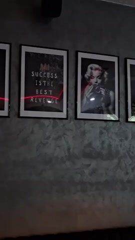 The video shows a wall decorated with framed pictures and a sculpture. The pictures are in black and white and appear to be of Marilyn Monroe, a cityscape, and a vehicle. The sculpture is of a gorilla in a colorful, drip-painted style. A red laser beam is visible crossing the image. The wall has a textured finish. A pendant light fixture hangs from the ceiling.