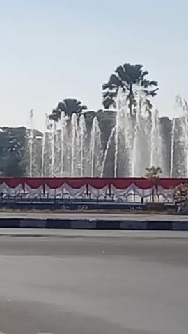 The video captures a street scene with a fountain in the background, decorated with red and white banners. Several vehicles are visible, including a dark-colored car in the foreground, a white van with a license plate that reads "DR 7443 AS", and a motorcycle with two riders. Another motorcycle is seen from behind, with one rider wearing a dark top and the other a yellow hooded sweatshirt. The environment appears to be outdoors, possibly in a tropical location, given the presence of palm trees. The sky is clear and blue, suggesting daytime. The overall atmosphere is that of a busy intersection or roundabout.