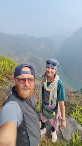 A man and a woman are taking a selfie against a mountainous backdrop. The man, on the left, is wearing a blue baseball cap, glasses, and a gray vest. He has a reddish beard and is smiling. The woman, to his right, has a blue patterned headscarf, sunglasses, and a scarf, and she's wearing a green t-shirt with a plaid shirt around her waist and a crossbody bag. They are standing on a rocky area with green vegetation. A river is visible in the valley below. The woman raises her arms. The man gives a thumbs-up.