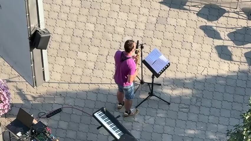 A person plays the saxophone while standing on a paved area. The musician is wearing a purple t-shirt and denim shorts. In front of them is a music stand with sheet music, and to their left is a keyboard. The scene appears to be outdoors, with sunlight casting shadows on the ground, suggesting it is daytime. There is a speaker mounted on a wall or structure to the left of the frame, and some chairs are visible in the background. The paved area has a patterned design. The general atmosphere seems to be one of a casual outdoor performance or practice session. The location is identified as Slobozhanske, Ukraine. There is no visible text in the frame.