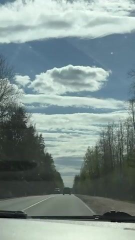 The video shows a view from inside a moving car looking out the windshield. The car is on a two-lane highway that stretches into the distance, bordered by tall trees on both sides. Two cars are visible ahead in the same lane. The sky above is blue with layered, elongated clouds. The car's dashboard and windshield wipers are visible at the bottom of the frame. Two people are talking.