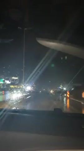 A night-time view from the dashboard of a moving car shows a wet road and street lights reflected on the wet ground. There are buildings with lights in the distance. The scene is lit only by streetlights and car headlights. The windshield wipers are not active and drops of water are visible on the windshield. The dashboard is black. The inside of the car is dark except for a bit of reflection on the dashboard.  The car is driving slowly. Music plays in the background.