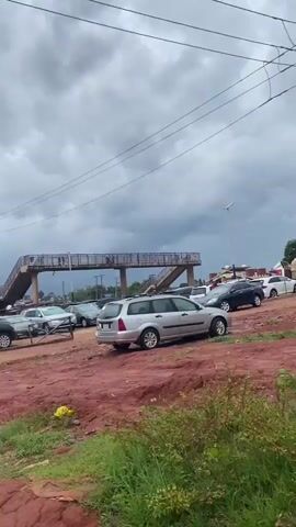 The video captures an outdoor scene in Auchi, Nigeria, dominated by a large parking area with reddish-brown laterite soil. The ground is uneven, with visible wet patches suggesting recent rain or damp conditions.

The recording begins with a close-up view of the rear of a silver Ford Focus station wagon, which is parked facing away from the camera. The camera then slowly pans left, revealing a broader expanse of the parking lot filled with numerous parked vehicles of various makes and models, including several silver, black, and white sedans and SUVs. These cars are parked somewhat haphazardly on the unpaved surface.

In the midground and background, a prominent metal pedestrian bridge with concrete supports and stairs spans horizontally across the frame. Several individuals are visible walking across the bridge, indicating it's in active use. Overhead, power lines crisscross against a heavily overcast and cloudy sky, suggesting dull weather conditions. The overall setting appears to be a busy outdoor area, possibly a market, transport hub, or car lot, given the concentration of vehicles and the pedestrian infrastructure. No discernible text is visible on any signs or vehicles.