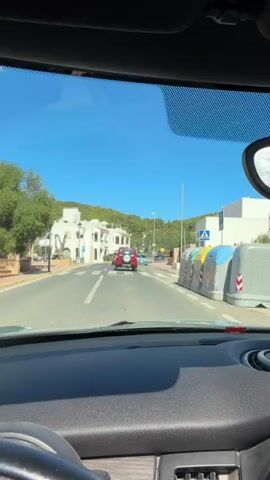 The video is taken from inside a car, looking through the windshield. The car is moving down a street in what appears to be a town or village. There are buildings on both sides of the street, many of them white with Mediterranean architectural styles. Trees and green hills are visible in the background under a clear blue sky. Several cars are on the road, including a red SUV in front of the car the video is being filmed from, and a blue car further ahead. There are also streetlights, trash bins, and pedestrian crossings visible. The interior of the car shows part of the dashboard and steering wheel. The overall impression is a sunny day drive through a picturesque town.