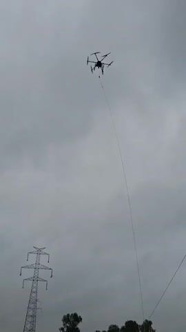 A drone is flying in the sky, tethered to the ground by a long, thin cable. Below the drone, a person wearing a high-visibility vest is standing in a field, holding the cable. Several large spools of cable are on the ground near the person. In the background, there are trees and a large metal electrical tower. The sky is overcast.
