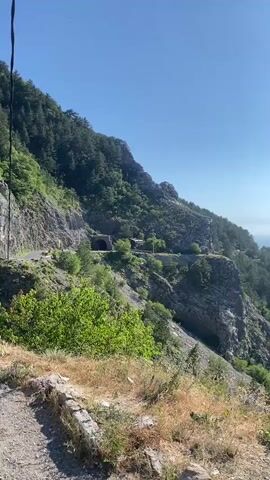The video pans across a steep, rocky mountainside covered in lush green trees and brush. A winding road is visible carved into the side of the mountain, with a tunnel entrance appearing halfway up. The sky is clear and blue, with the sun glinting off the water in the distance, which appears to be a bay or a large lake surrounded by more mountains. The foreground is a dirt path with dry grass and some low-lying vegetation.