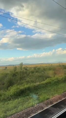 The video shows a view from a moving train window looking out onto a rural landscape. The sky is blue with scattered white clouds. The landscape is mostly green fields and bushes, with some trees in the distance. There are power lines visible across the top of the frame. In the foreground, the train tracks and the side of the train car are visible.