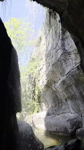 The video shows a view from inside a cave looking out at a waterfall. Water streams down the rocky wall opposite the cave entrance, feeding into a small pool. Beyond the waterfall, green foliage and a tree are visible under a bright, blue sky. The cave is dark and damp, with water cascading down the entrance.