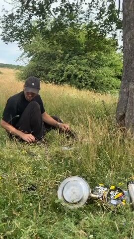 A young person wearing a black t-shirt, black pants, and a black baseball cap is crouched down in a field of tall grass next to a large tree. They are looking down and appear to be interacting with something in the grass in front of them. To the right of the person, on the ground, are several objects, including what appears to be a metal pot, some coiled wire, and a few yellow and black items. The background shows more grassy fields and trees under an overcast sky.
