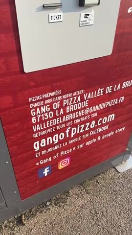 The video begins with a close-up of a red, textured surface with white text. The text indicates that pizzas are prepared daily in their workshop, provides the business name "GANG OF PIZZA VALLÉE DE LA BROQUE", an address and postal code, a website address "gangofpizza.com", an email address, and social media handles (Facebook, Instagram, Apple, and Google). The camera then pans up to a white panel with various slots and buttons, including a ticket dispenser, a card reader, and a keypad. A close-up shot follows of a digital screen displaying "Product(s) remaining to be delivered: 1/1" and "Your Margarita is on its way...". The background of the screen shows a blurred image that appears to be a hand reaching for something. The overall scene depicts a pizza vending machine and its interface, suggesting a transaction has occurred or is in progress.