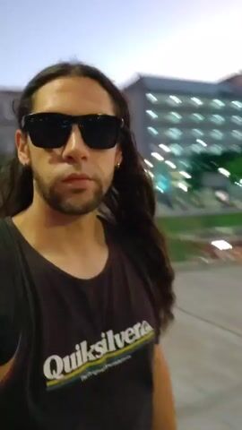 A man with long hair and sunglasses records a video of himself in a public space. He wears a black tank top with a "Quiksilver" logo. In the background, a group of people is exercising in a plaza with surrounding buildings and trees. The scene appears to be at dusk or twilight.
