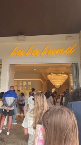 A crowd of people are waiting in line to enter a store called "La La Land." The store has a bright interior, with a large, tiered chandelier hanging from the ceiling. The lighting inside is warm and inviting. People are chatting and some are holding drinks or other items, presumably purchased from the store. The camera angle suggests the viewer is also standing in line, looking towards the entrance.
