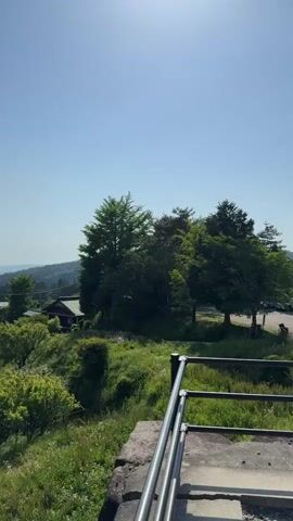 The video shows a scenic overlook of a Japanese countryside. From a slightly elevated position behind a silver railing, the camera pans slowly left to reveal a winding road lined with traditional Japanese houses, lush green trees, and rolling hills in the distance. The sky is a clear blue with a few scattered clouds. Two motorcycles can be seen traveling along the road. The landscape suggests a peaceful, rural setting in Japan.
