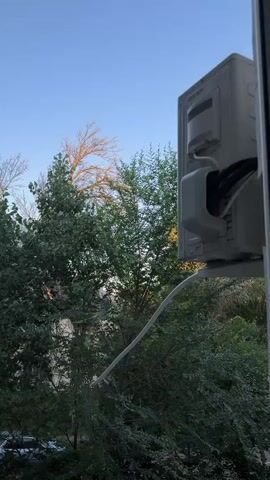 The video captures an outdoor unit of an air conditioner mounted on a white bracket outside a window. The unit is white and shows electrical wires connecting to it. A white pipe extends from the unit and curves downwards. Behind the air conditioner, lush green trees fill the background. The sky above is a clear blue, with a hint of warm light illuminating the branches of a bare tree in the distance, suggesting it may be sunrise or sunset. The scene appears to be from an urban or suburban setting due to the presence of buildings and trees. No people are visible in the video.