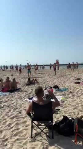 A sunny beach scene depicts a group of shirtless young men playing with multiple soccer balls in the sand.  The men, wearing various swim trunks, are spread out in a loose formation kicking and heading the balls. To the foreground, several people are seated or lying on the beach, observing the game. A man sits in a folding chair with his back to the camera. Beach towels and bags are scattered around the foreground. The sky is clear and blue, and the ocean stretches out to the horizon with a breakwater in the distance. The beach is covered in light-colored sand.