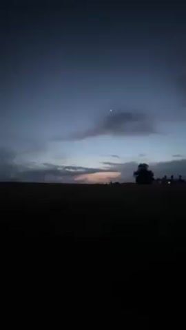 The video depicts a dark, rural scene with a road stretching into the distance under a cloudy twilight sky. There's a crescent moon visible in the sky. In the foreground, a dark pickup truck is parked on the side of the road, with at least two people standing nearby. Lights from distant buildings can be seen along the horizon. The overall atmosphere is dark and somewhat desolate.