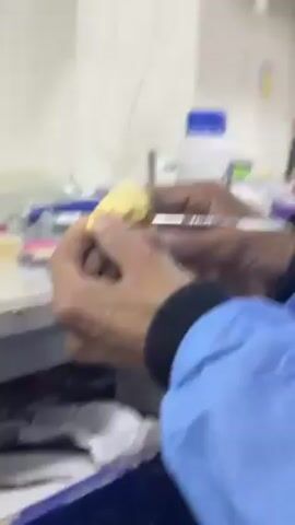 In a sterile, indoor environment likely a dental lab, a person wearing a light blue lab coat is meticulously working on a yellowish dental mold. The person is using a metallic dental instrument to carefully sculpt or adjust the mold. The workbench is littered with dental tools and materials, including a bottle of liquid, a dental drill, and various dental pieces. The overall focus is on the precision and detail required for dental work.