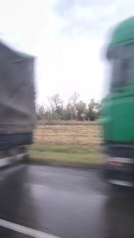 The video captures a view from inside a moving vehicle, likely a car. The scene outside the vehicle is blurry due to motion, showing a rural landscape with fields and trees under an overcast sky. The interior of the vehicle is visible, including a seat and a window. The time stamp indicates the video is a few seconds long.