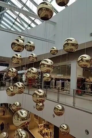 The video shows a wide shot of a shopping mall interior. The camera is looking up at a multi-story atrium. Suspended from the ceiling by thin wires are numerous large, reflective, spherical ornaments. These ornaments are gold or brass colored and distort reflections of the mall's interior, including the ceiling, lights, and the floors below with shops. The mall appears to be modern with clean lines and a glass roof. In the background, shops are visible on different levels, with displays and mannequins. No people are clearly visible or in focus. The video is static with no camera movement.