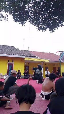 A person in dark clothing with their hair tied up is performing a martial arts move in a low crouch, with their right arm extended upwards and their left arm bent at the elbow. They are on a raised red carpeted area. Several other people are sitting and watching, some in the foreground and some in the background. Behind them, there is a yellow building with a red roof and a large tree with dark green leaves overhead. The sky is overcast. There is a tall pole with a wire extending from it in the background.
