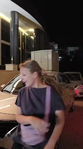 A young woman with long blonde hair pulled back in a ponytail, wearing a dark t-shirt and a pink strap across her body, stands in front of a light-colored sedan at night. She initially turns her back to the camera, then faces it, making a gesture with her hands as if to wave or motion. The car's headlights are on, and the background shows a modern building with illuminated windows and dark columns. The scene is lit by artificial lights, creating a night-time ambiance.