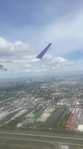 The video shows an aerial view of a city from an airplane window. The plane's wing with a blue tip is visible in the upper left corner of the frame. Below, a sprawling urban landscape extends to the horizon, consisting of buildings, roads, and patches of greenery. The sky is blue with scattered white clouds. The overall tone is calm and picturesque.