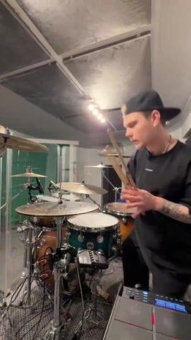 The video shows a man playing drums in a music studio. He is dressed in a black t-shirt and black cap, and he is seated behind a drum set with multiple cymbals and drums. His arms are visibly tattooed. The drum set is behind a clear, possibly plexiglass, sound barrier. The room is well-lit with what appears to be a strip of lights above, and the ceiling has a textured, sound-dampening design. A electronic drum pad is situated next to the drum kit. The man throws his drumsticks in the air and catches them during his performance.