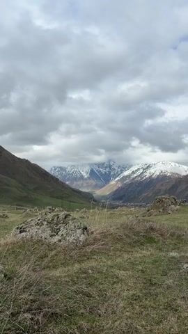 The video shows a mountainous landscape under a cloudy sky. The foreground features a grassy field with scattered rocks. In the midground, a valley stretches between two green mountain slopes, leading to a towering mountain range in the background. The peaks of the mountains are covered in snow and partially obscured by clouds. A small structure is visible in the valley. Overhead power lines cut across the landscape.