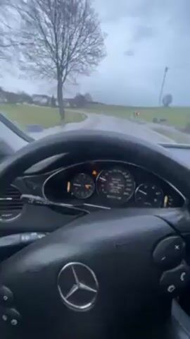The video shows the dashboard and steering wheel of a Mercedes-Benz car, with the driver's perspective of the road ahead. The car is driving on a wet road, likely in a rural area, with green fields and some houses visible in the background. The sky is overcast, suggesting a cloudy or rainy day. The steering wheel is black with the Mercedes-Benz logo in the center. The dashboard features the speedometer, fuel gauge, and other instrument dials. The car appears to be driving slowly, and the dashboard lights are illuminated. The music is playing in the car.
