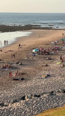 A wide shot captures a crowded sandy beach with a rough ocean in the background. The beach is populated with numerous people, many of whom are lying on towels or sunbathing. Several colorful umbrellas are dotted across the sand, providing shade. Some individuals are standing or walking, while others appear to be engaged in conversations or activities. The ocean water is a deep blue, with visible waves breaking near the shore and rocky outcrops. The sand is a light brown, with a textured surface. In the foreground, large rocks are scattered, and a grassy incline is visible to the right. The overall lighting suggests a bright, sunny day, likely late afternoon or early evening, given the soft sunlight.