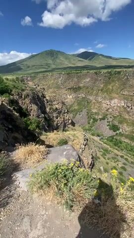 This is a vertical video of a canyon in Artashavan, Armenia. The video pans from right to left, showing a vast canyon with steep, rocky walls covered in green vegetation. The canyon floor is lush with trees and appears to have a river or stream running through it. In the foreground, there are large rocks and dry grass, with yellow wildflowers blooming. The sky is a bright blue with scattered white clouds, suggesting it is a sunny day. In the distance, rolling green hills and mountains are visible, and a faint hazy outline of a town or city can be seen on the horizon. The overall impression is one of natural beauty and grandeur, with a sense of scale and vastness. The video is likely taken from a viewpoint overlooking the canyon, possibly a hiking trail or scenic overlook. The time of day appears to be midday or early afternoon due to the bright sunlight and clear sky. There are no people or specific objects clearly visible besides the natural landscape, though a shadow of a person taking the video can be seen in the bottom right corner. The mood is peaceful and awe-inspiring.