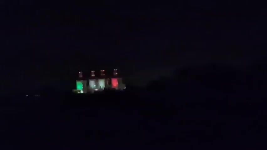 The video begins with a view of a dark, outdoor environment at night. A building, possibly a power station or industrial facility, is visible in the distance, illuminated by red and green lights. The camera appears to be mounted inside a moving vehicle, as evidenced by a faint barrier in the foreground and occasional bright lights from oncoming traffic passing by. The building's lights are the primary source of illumination and are the only clearly distinguishable features in the frame. The video progresses with the vehicle continuing its movement, causing the building to become more or less visible as it moves in and out of frame or as the vehicle's trajectory changes. There are no people or other identifiable objects or actions depicted.