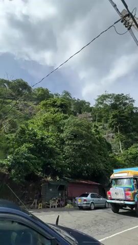 The video shows a colorful truck driving on a road next to a hillside with lush green trees. The truck has a red cab, a blue and yellow striped container, and the word "NT" on the back. The sun is shining brightly through the clouds, creating a slightly hazy effect. There are power lines visible above the road. In the foreground, part of a dark blue car is visible. On the side of the road, there is a small structure resembling a shop, and people are seen standing nearby. There is also a three-wheeled vehicle parked by the side of the road, and a person standing near it. The overall setting appears to be a rural or semi-rural area with a hilly landscape.