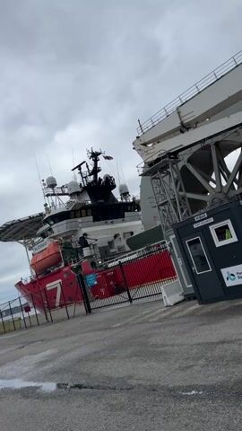 The video shows a dock with a large ship moored alongside, a port security booth, and a massive crane.  The ship is red and white, with a helicopter landing pad on the stern. The port security booth is grey and black, with “HIBAS PORT SECURITY” printed on the side. The crane is grey and white, with the word “subsea” visible on the side. The sky is overcast and grey. There are two people on a platform next to the ship. A trash can is near the port security booth. The camera pans to the right.