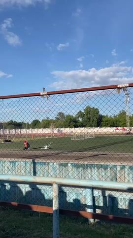 The video shows a speedway race from behind a chain-link fence. Several motorcycles are racing on a dirt track. One person in a yellow vest appears to be an official. The sky is blue with some clouds, and there are trees and buildings in the background. The fence in the foreground is rusty.