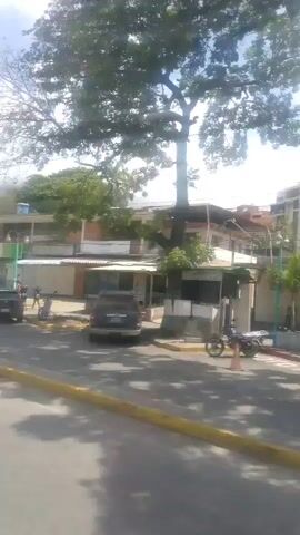 The video shows a street scene with buildings and trees. There are cars parked on the side of the road and people walking. One person appears to be cleaning the street. The sky is cloudy. The video is shaky, suggesting it was filmed from a moving vehicle.