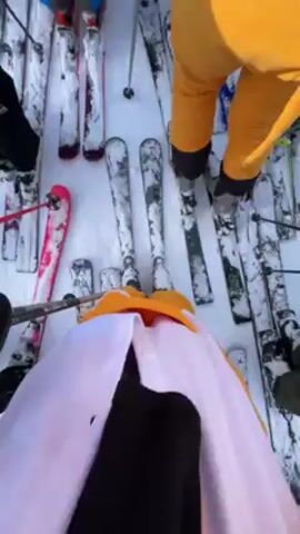 The video shows a group of people standing on skis in a snowy environment, possibly a ski resort. The person filming is wearing a yellow and white costume, possibly a cartoon character. Other individuals are wearing ski gear, including helmets and jackets. A mountain is visible in the background. The person filming turns the camera to show their face, smiling at the camera. The scene suggests a social gathering or group outing for skiing.