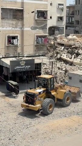 The video shows a street scene with damaged buildings and a yellow front-end loader. In the foreground, a group of people are gathered around a water truck, filling containers with water. There are many empty plastic containers and jerrycans visible. The people appear to be from a variety of ages, including children and adults. Some people are kneeling to fill their containers, while others are standing and waiting. The buildings in the background show signs of damage, with visible bullet holes and debris. A small shop with clothes displayed outside is also visible. The ground is covered in gravel and dirt. The overall impression is one of hardship and the necessity of resourcefulness.