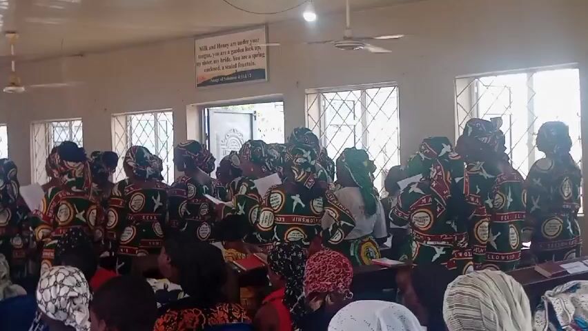 The video shows a group of people singing in what appears to be a church. The main focus is on a choir consisting mostly of women wearing matching patterned outfits and headwraps, holding songbooks. They stand facing the congregation. Some members of the congregation are visible in the foreground, seated and looking towards the choir. A person with a microphone appears to be directing the choir. The room has several windows providing natural light, and there are ceiling fans and fluorescent lighting fixtures. Two framed signs hang on the wall with scriptural verses. The overall atmosphere seems to be one of religious singing and participation.