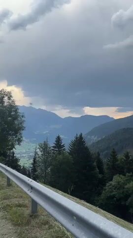 This media file captures a sweeping vista of a mountainous landscape, likely in the Freienfeld - Campo di Trens, Italy region. The scene is dominated by rolling green hills densely covered with coniferous trees, creating a lush, textured carpet that stretches across the slopes. In the distance, several layers of mountains fade into a hazy blue, hinting at a vast, expansive terrain.

The sky is a dramatic canvas of dark, heavy clouds, suggesting overcast or stormy weather. However, a sliver of bright, warm light breaks through the clouds on the horizon, casting a soft glow and hinting at the setting sun or a break in the weather. This contrast between the dark clouds and the warm light adds a sense of depth and mood to the scene.

In the foreground, a silver guardrail runs along a road or pathway, indicating the viewpoint is from an elevated position, likely a road winding through the mountains. The guardrail and the grassy verge next to it are clearly visible, grounding the viewer in the immediate environment.

There are no people or distinct objects, other than the guardrail, visible in the media. The activity is purely observational, showcasing the natural beauty of the mountainous region. The time of day appears to be late afternoon or early evening, given the quality of the light. The overall emotion evoked is one of awe at the grandeur of nature, with a touch of melancholy or anticipation due to the dramatic sky. No visible text is present in the media.
